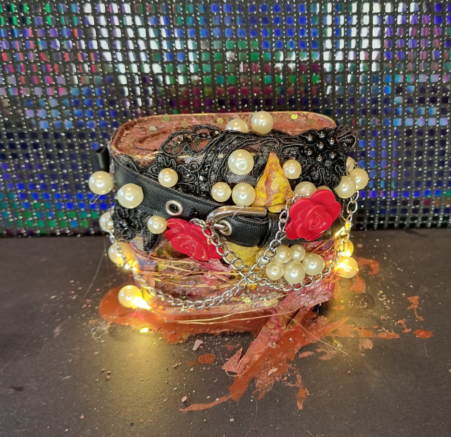 A cylindrical red-brown object is wrapped in black lace-like material, a belt and silver chains, with pale beads, red rose ornaments and glowing warm lights.